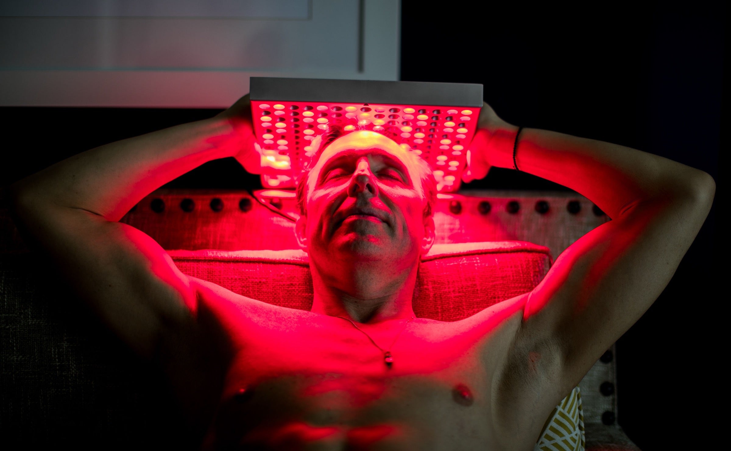Dave Asprey reclining with eyes closed, holding a TrueLight red light panel overhead to bathe his face and chest in red light